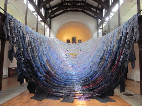Large textile installation made of layered, colored fabric strips hanging in a curved formation inside a hall with wooden floors and arched ceiling, part of an art exhibition Lahore.