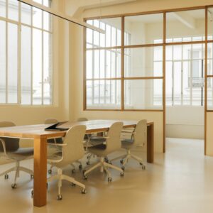 Modern industrial office space featuring a large wooden meeting table with six light gray ergonomic chairs on casters, surrounded by large windows and glass partition walls allowing natural light to fill the room.